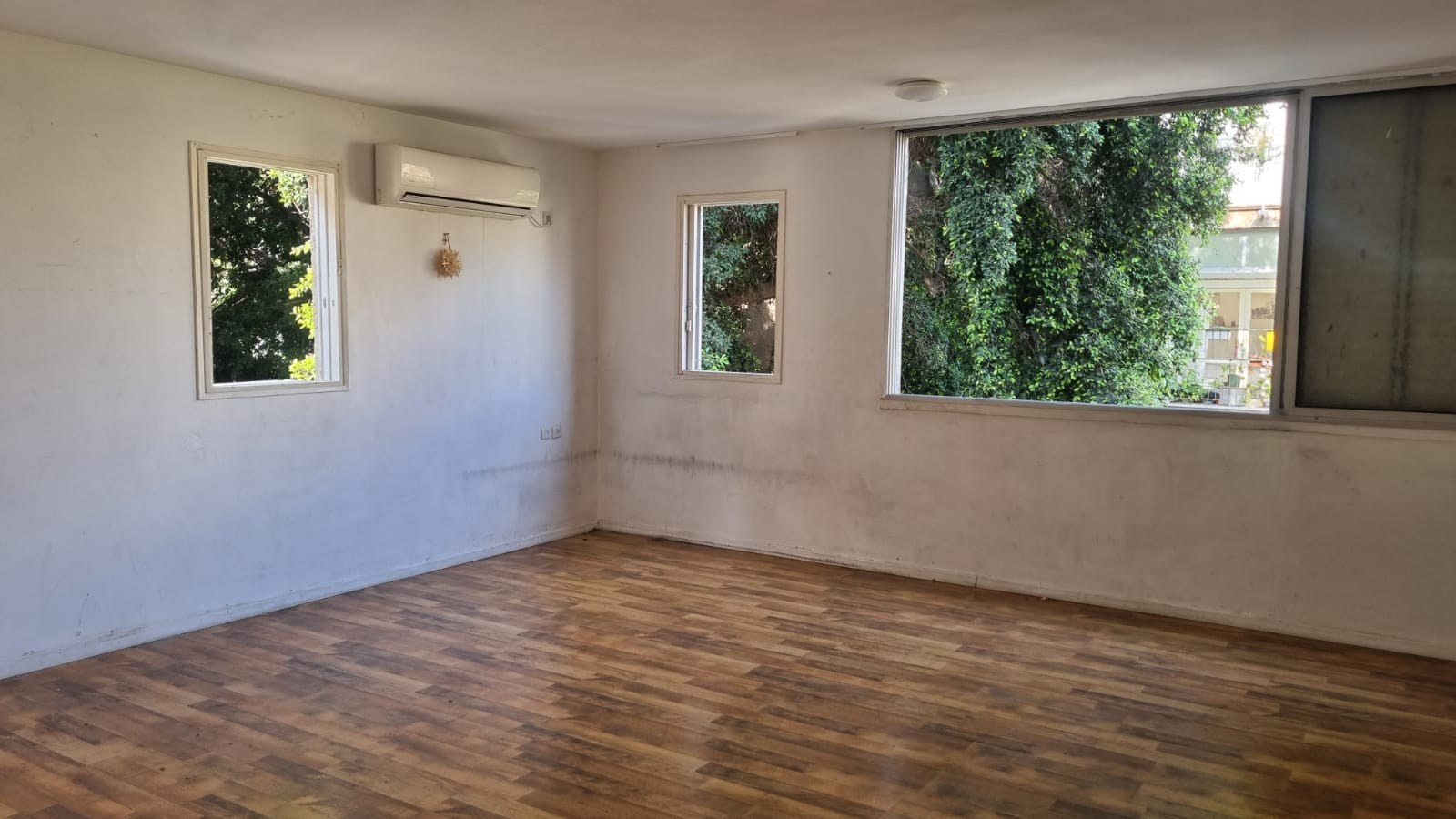 Empty living room with wood-laminate flooring, white walls, split-system AC, and three windows overlooking dense greenery