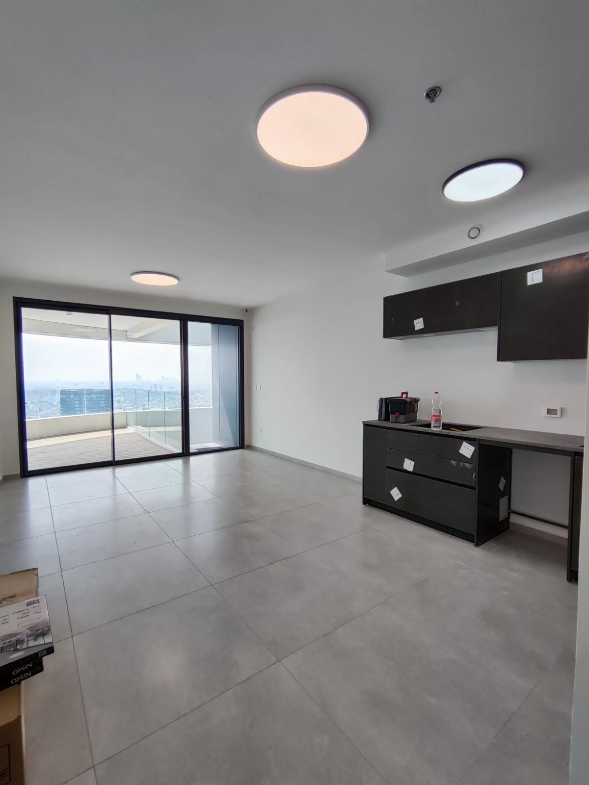 Open-plan living area with dark charcoal kitchenette, large-format gray floor tiles, and black-framed sliding doors to a high-floor balcony with city views
