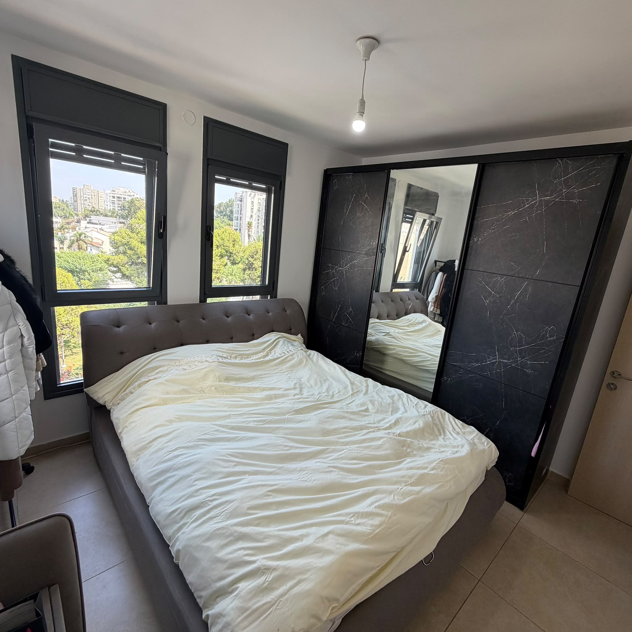 Modern bedroom with gray tufted bed, marble-pattern sliding wardrobe with mirror panel, and black-framed windows over trees and city buildings